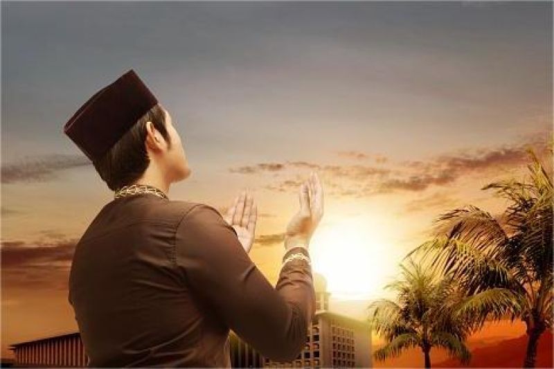 tangan seseorang mengangkat doa di langit senja dengan latar belakang masjid